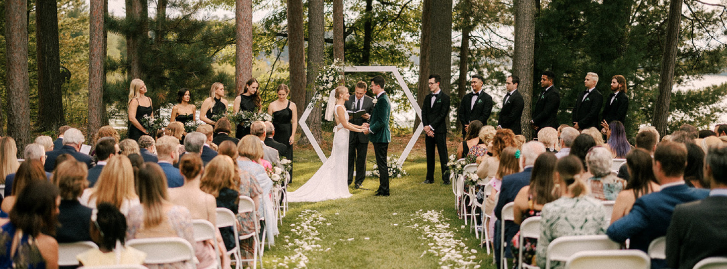 bride and groom exchanging vows in front of a white structure in a pine tree forest overlooking Lake Manitowish with a crowd of witnesses and white rose petals strewn along the aisle.