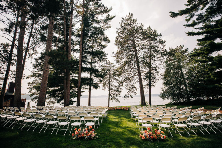 wedding ceremony setup. white chairs on green lawn with tall pine trees and lake in the distance. bright colored flowers on the ground at the entrance to the aisle and at the end.