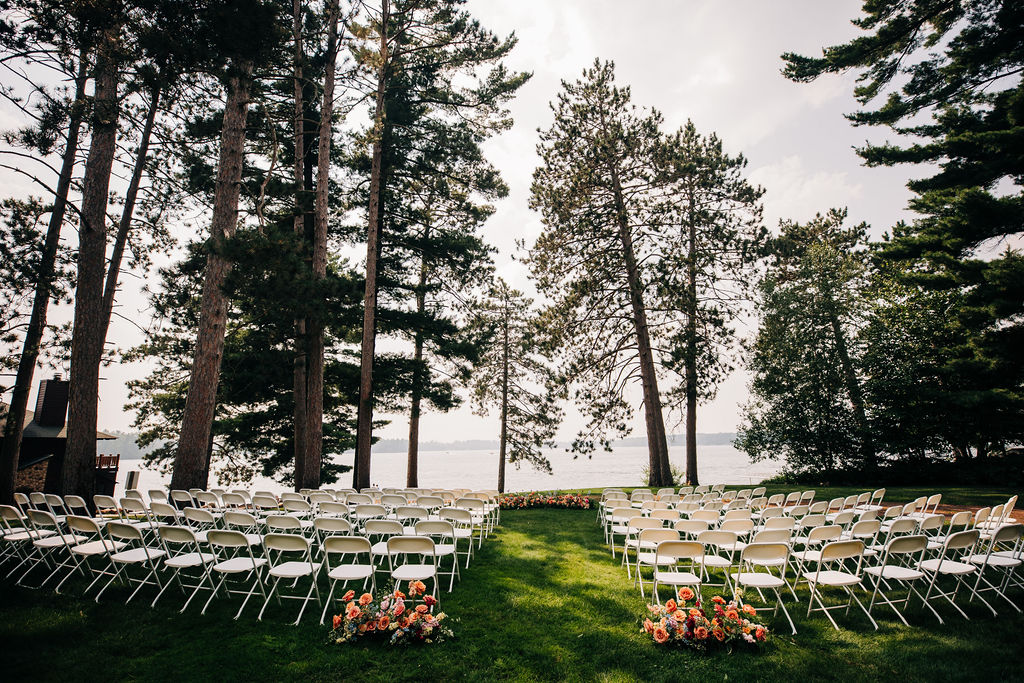 wedding ceremony setup. white chairs on green lawn with tall pine trees and lake in the distance. bright colored flowers on the ground at the entrance to the aisle and at the end.