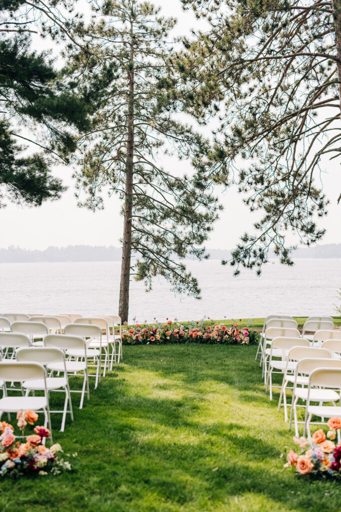 lakeside wedding ceremony setup with a main aisle on a green lawn. white chairs on either side of the aisle and at the end of the aisle under pine trees and before the lake, a hedge of multi-colored floral arrangements on the Lakeside Lawn at Chippewa Retreat Resort in Manitowish Waters, WI