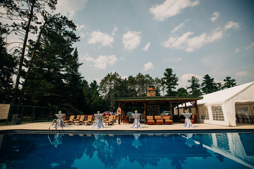 pool in foreground with pine trees, blue sky with clouds, exterior structure with lounge chairs by the pool and a white tent on the far right side of the image.