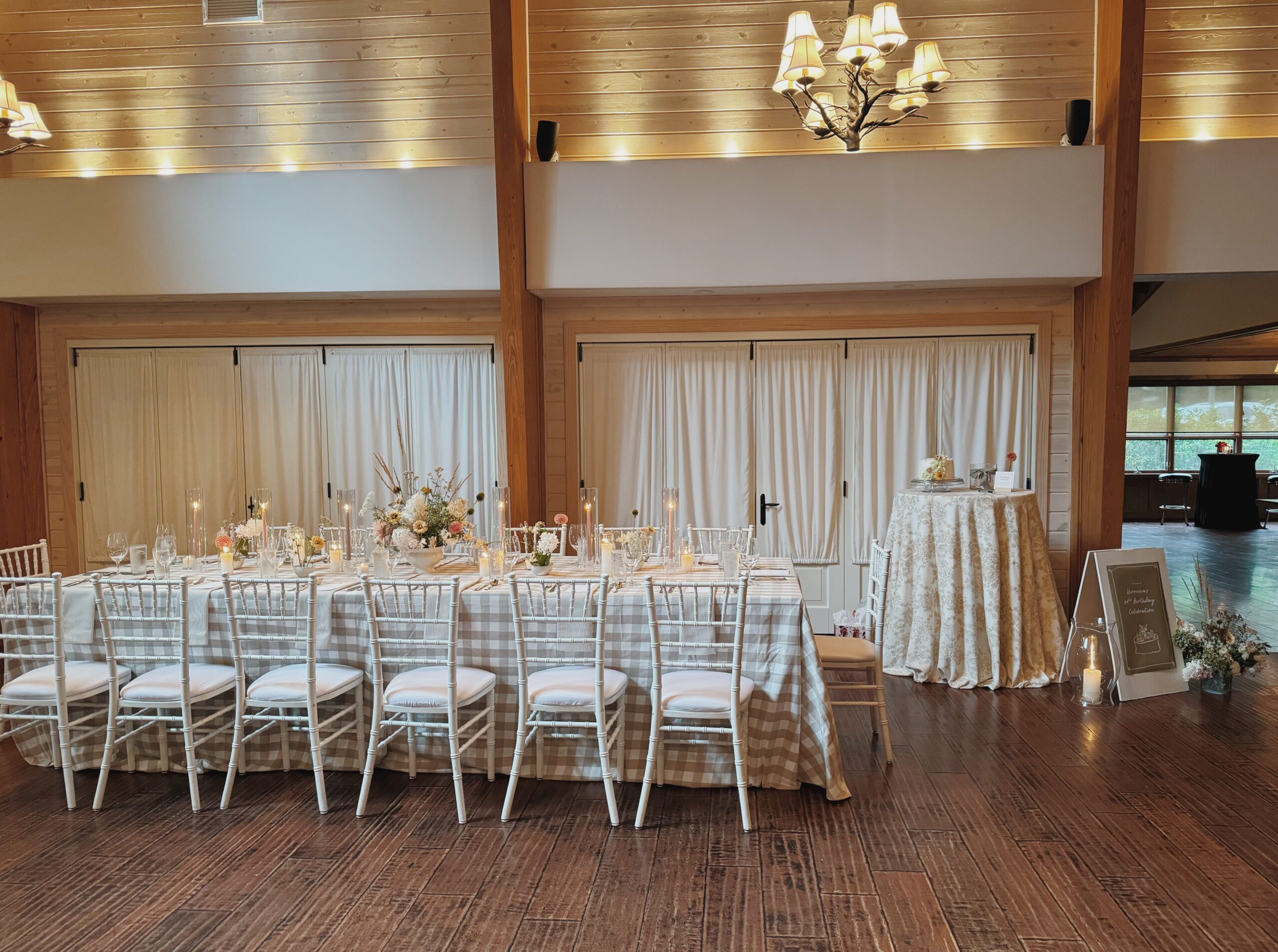 wedding reception table set with a tall draped cake table on the right side. Set in a high-ceiling, lodge style reception room with brand-style chandeliers.