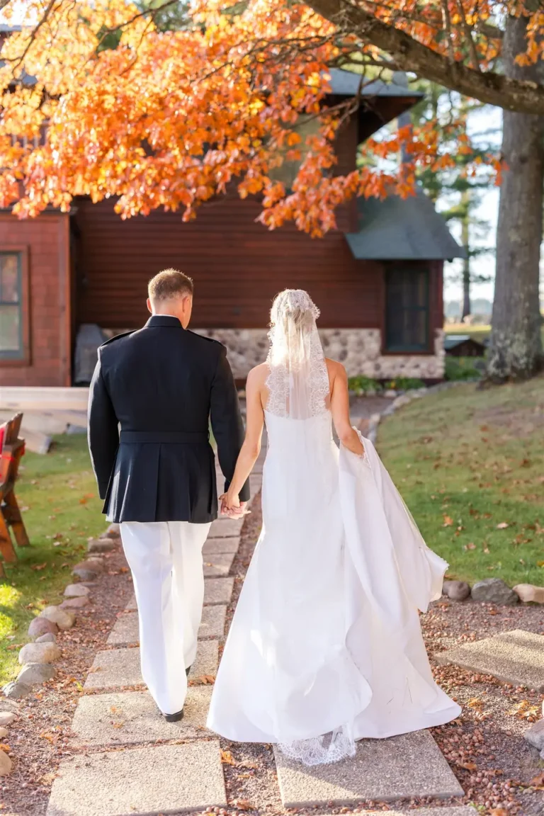 Bride and Groom walking down gravel and paved path towards a wood-sided building. Trees with orange leaaves above their heads.