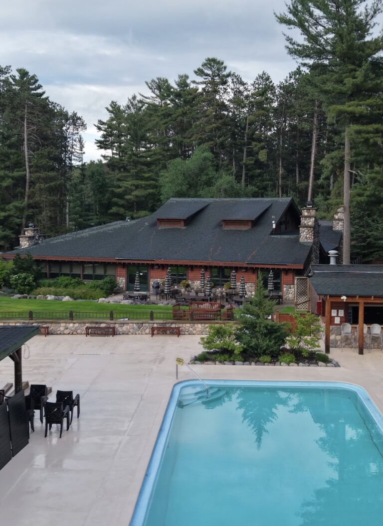 Aerial picture of the a pool and cabana with a glass wall surrounding on a grassy hill with a lake to the left