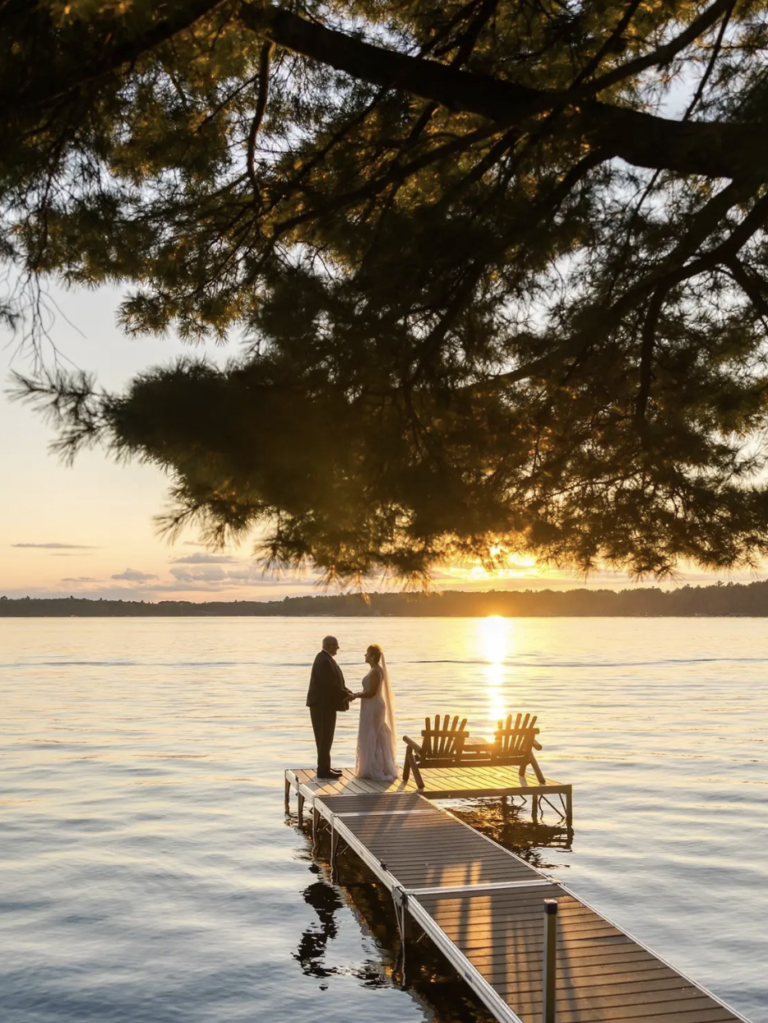 Bride and Groom standing on dock on a lake with pine trees extending over their heads as the sun sets in the distance