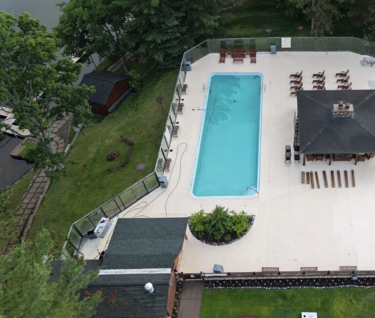 Aerial picture of the a pool and cabana with a glass wall surrounding on a grassy hill with a lake to the left
