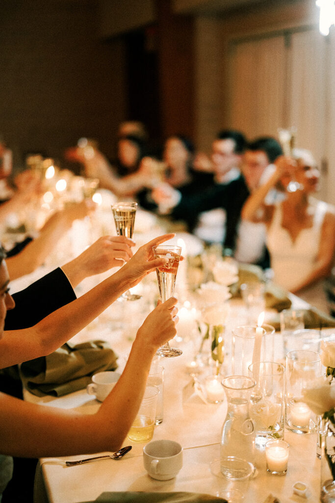 candlelit wedding reception dinner with guests and bride and groom raising cocktails and champagne glasses in the air for a toast over a white table cloth table with white roses