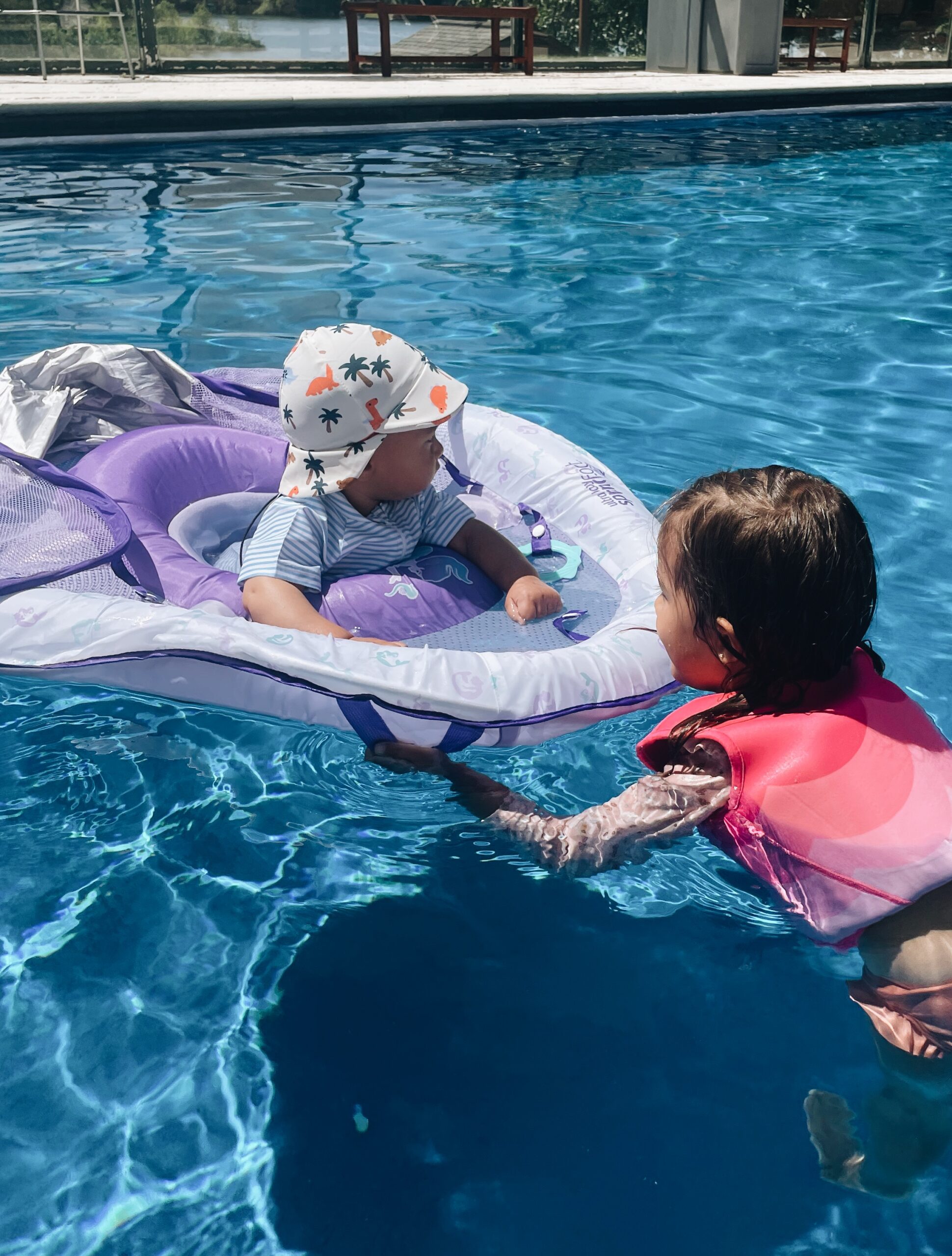kids swimming in pool together. baby floating in purple raft, little girl in bright pink pool vest