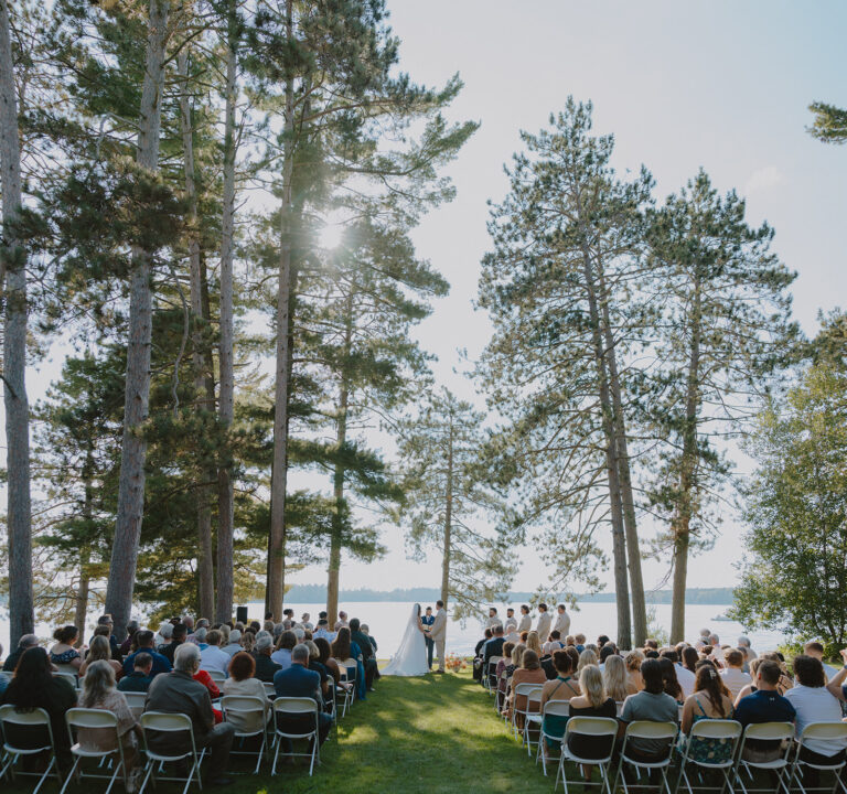bride and groom getting married with a crowd of people sitting in chairs on the grassy lawn. couple standing under pine trees in front of Lake Manitowish at Chippewa Retreat Resort in Manitowish Waters, WI