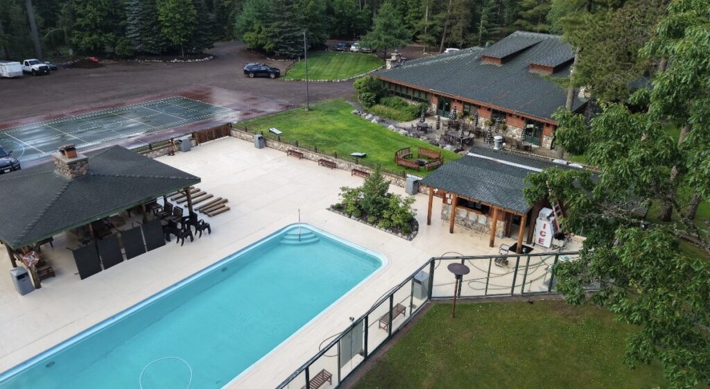 Aerial picture of the a pool and cabana with a glass wall surrounding on a grassy hill with a lake to the left