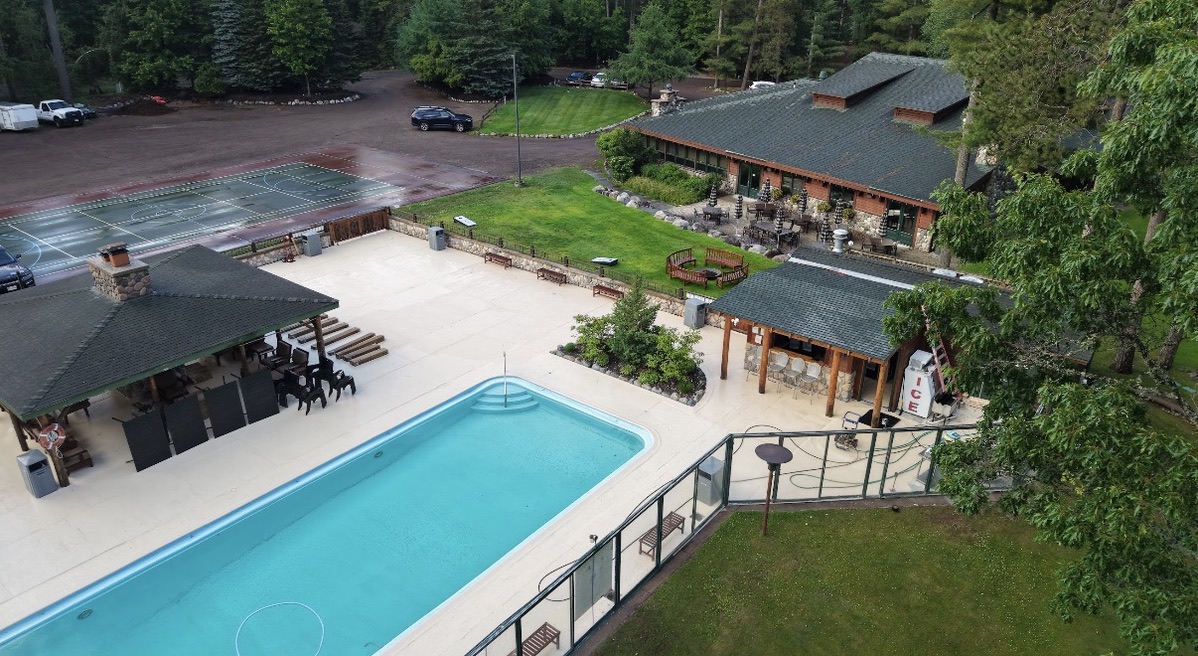 Aerial picture of the a pool and cabana with a glass wall surrounding on a grassy hill with a lake to the left