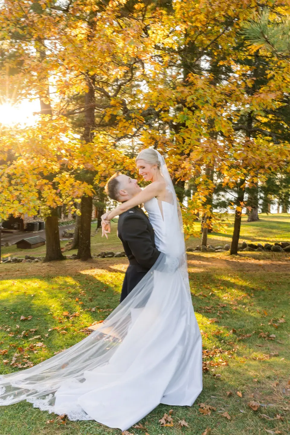 groom lifting bride up in the air. groom and bride kissing. standing on a green lawn with fall color trees behind them.