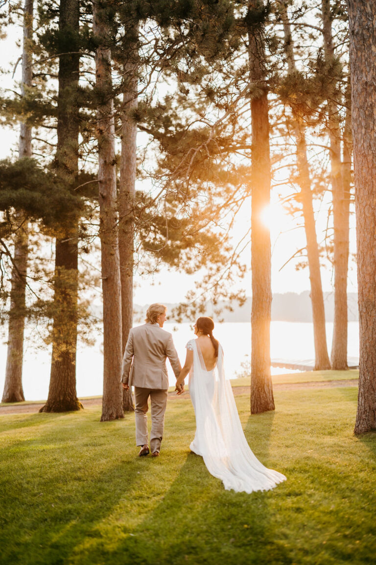 bride and groom with backs to the camera walking on a grassy lawn dotted with pine trees, walking towards a lake as the sun sets in the distance beyond them. Lakeside Lawn at Chippewa Retreat Resort in Manitowish Waters, WI