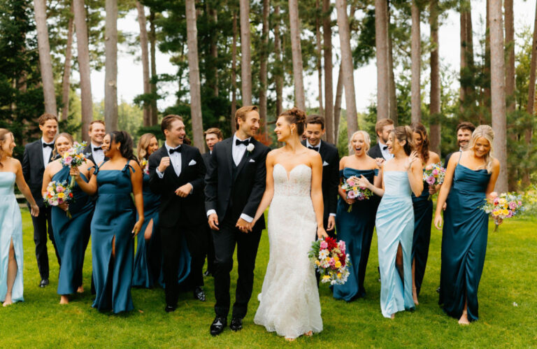Wedding party with bride and groom in front of them waking towards the camera excitedly. Gowns of blues. Men in tuxes. Pine trees behind them.