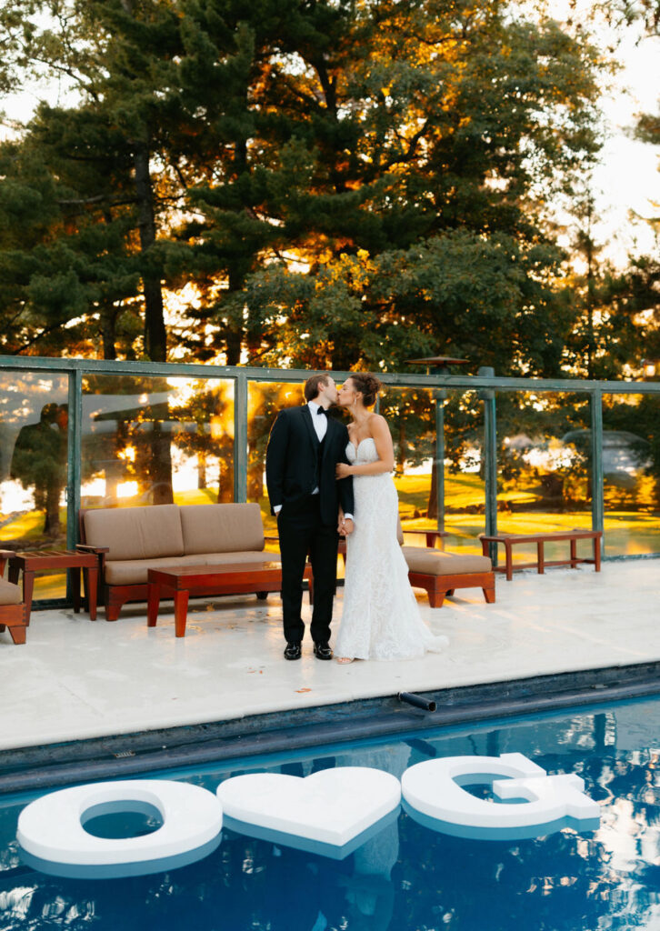 bride and groom kissing at the outdoor pool, pine trees behind and a big O-Heart-G sign floating on top of the pool water