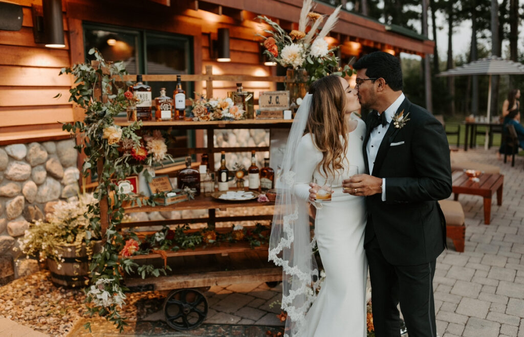 bride and groom kissing outside on reception patio with a vintage bar cart behind them with various whiskeys and cigars and flowers. each holding a glass of whiskey.