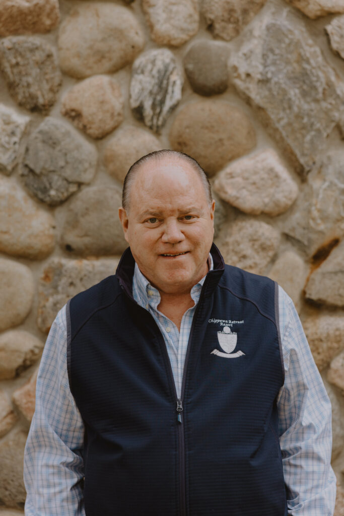 middle-aged man in a blue shirt and vest, smiling and standing in front of a stone wall