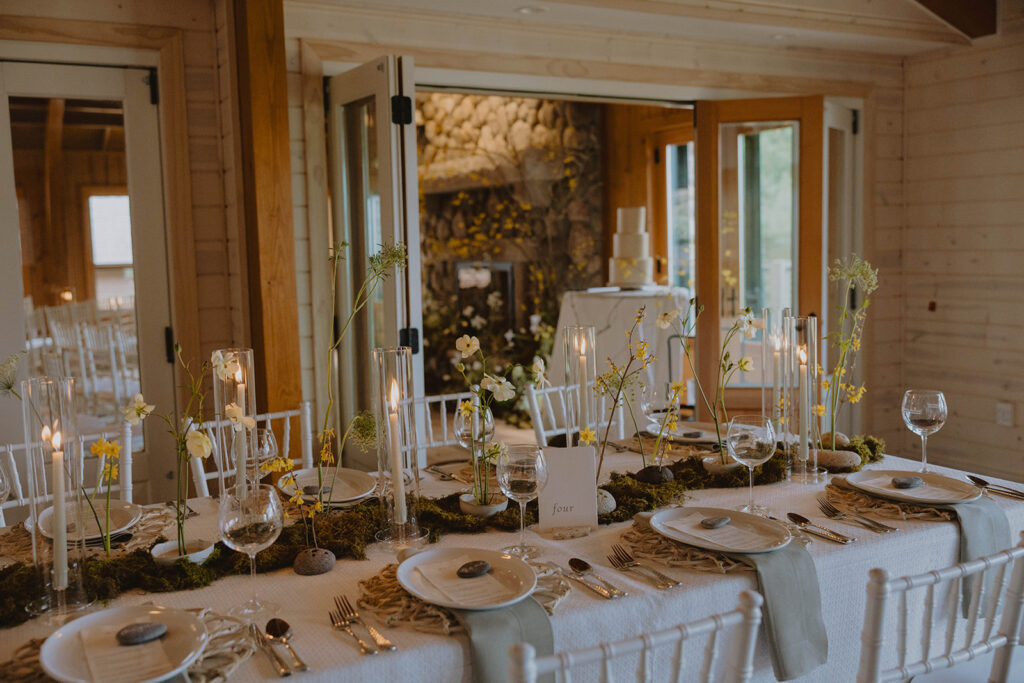 Set dining tables for a wedding reception with moss and daffodils and white tablecloths. a stone fireplace in the background with other floral decor. Micro-Wedding setup.