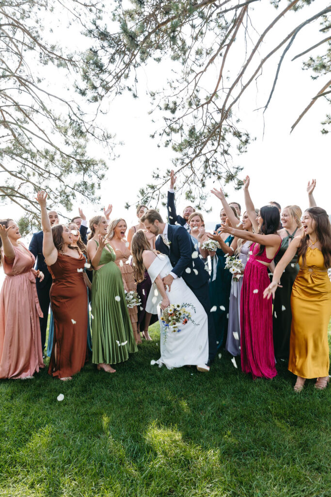 bride and groom kissing surrounded by bridesmaids and groomsmen in front of Lake Manitowish with pine trees behind them on a green grassy lawn at Chippewa Retreat Resort